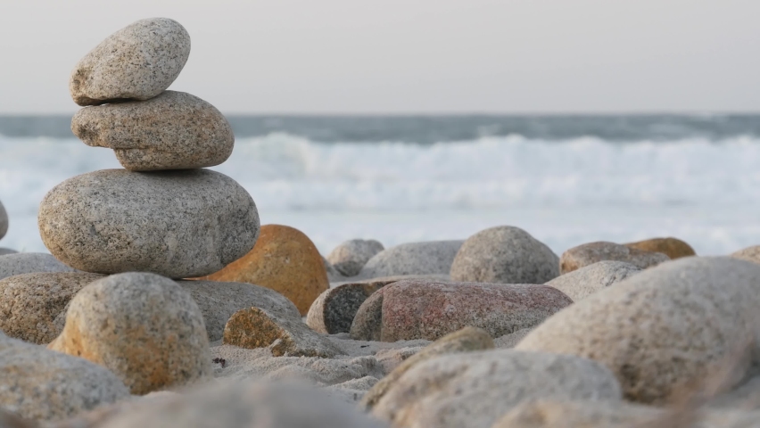 Rock balancing on pebble beach, Monterey 17-mile drive, California coast, USA. Stable pyramid stacks of round stones, sea ocean water waves crashing at sunset. Serenity harmony, calm zen meditation. | Shutterstock HD Video #1087016741