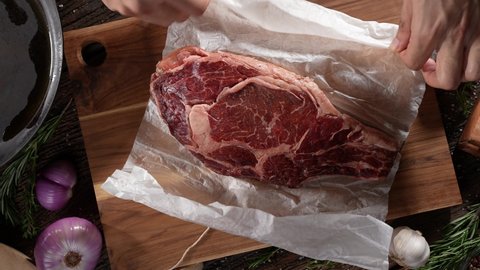 Unwrapping raw rib eye steak meat beef on wooden chopping board on a wooden table prepared for cooking. slow motion