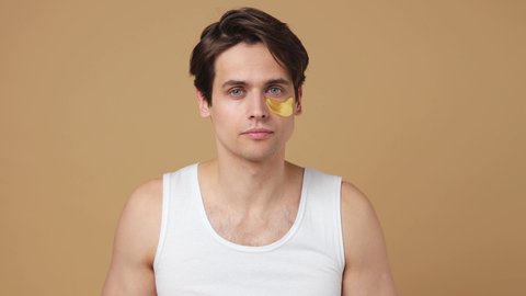 Satisfied young man 20s perfect skin wears white tank top put on wear gold patch under eye isolated on plain pastel beige background studio portrait. Body care healthcare cosmetic procedures concept