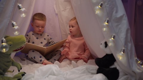 Brother and sister sitting in white tent wigwam quarrel with each other, fighting with their hands, fight between sister and brother, boy reading book for sister, sister slapping his brother hands.