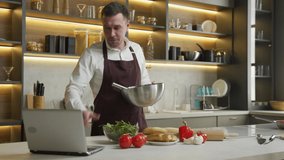 Young man in apron prepares dinner following video. Chef cooks dish with vegetables mixing sauce in bowl and watching recipe in laptop at table in kitchen