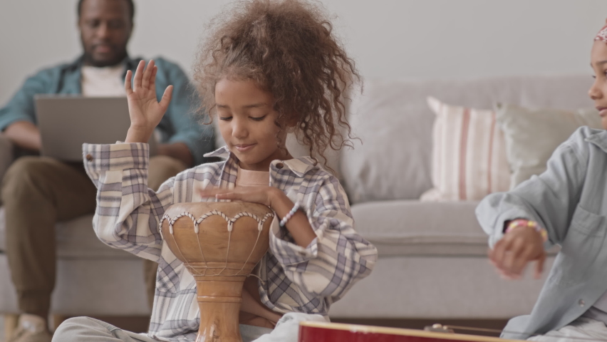 Handheld medium shot with slowmo of two pretty Biracial girls playing drum and guitar at home while their father working on laptop sitting on couch in background