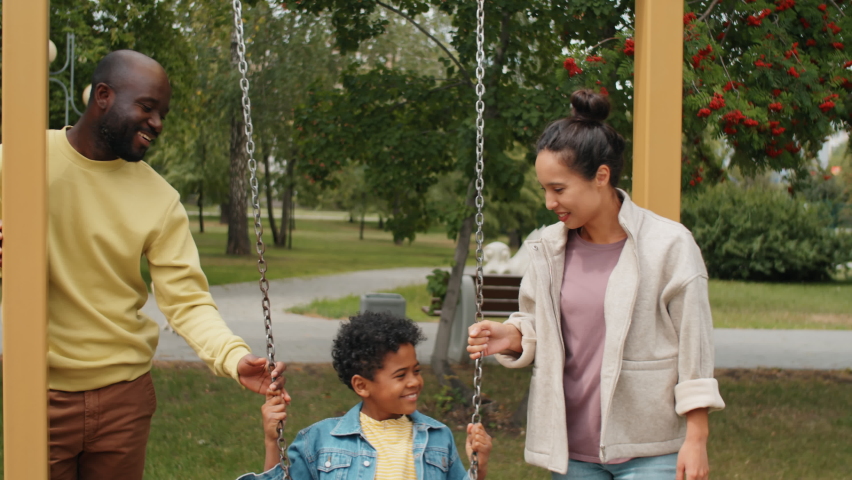 Little African American boy swinging on swing in park with help of happy mother and father