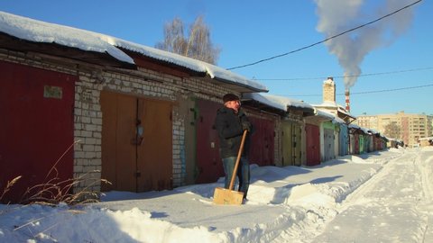 A man in warm clothes is leaning on a wooden shovel, cleansing the snow. Clear, sunny cool winter day, blue sky. There are snowdrifts all around. Climate change, city, there was a snowstorm. UHD 4K