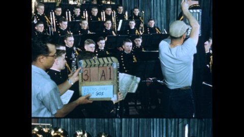 1950s: Men holding clapperboards, clapping. Conductor conducting orchestra. Orchestra performing. Musicians playing instruments.