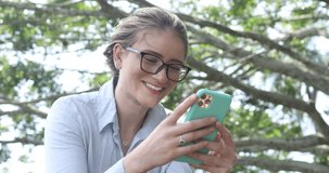 Young white woman using a mobile phone at day time with a green park in the background. High quality video. Mobile phone, technology, urban concept, smartphone. .