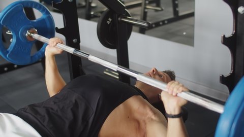 Male bodybuilder trains in gym with barbell in prone position on bench, front view. Athletic man lifts barbell at gym. Concept training and weightlifting