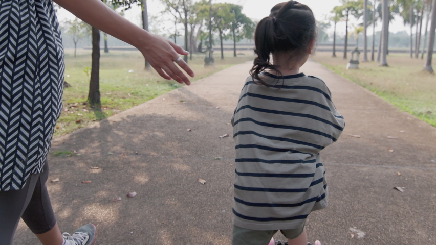 Asian kid girl is practicing scooter or roller board with her mother caring in beautiful park in the morning with sunshine. It shows leisure time activity of family for happily playing sport outdoor.