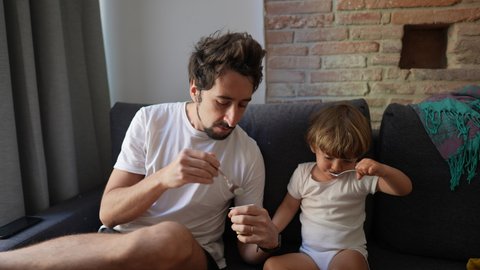 Father and toddler child eating breakfast together sitting in sofa at home