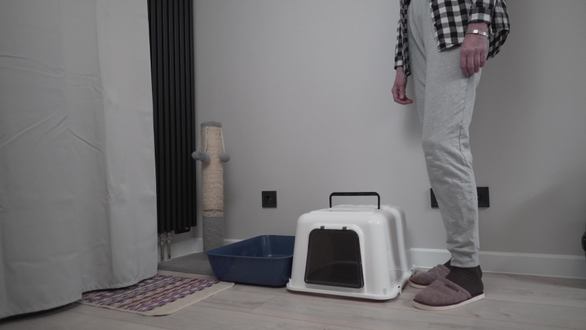 Senior woman assembles new cat litter box together with pet at home. Elderly female playing with cat sets hooded litter box for pet to go to toilet. Pet care, hygiene concept. Hooded cat litter pan