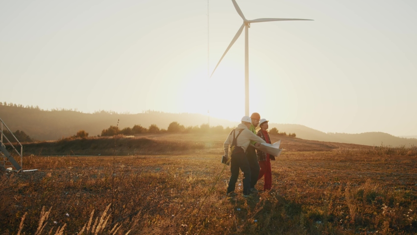 Engineers walking discussing blueprint in windmill farm inspecting wind turbine power station. Rotating at sunset. Slow motion Royalty-Free Stock Footage #1087047167