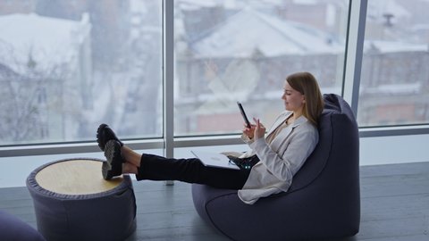Relaxed young woman sits in bean chair stretching her legs to the coffee table. Lady with laptop and glasses on her knees looks at her phone and smiles. Snowing weather backdrop in blur.