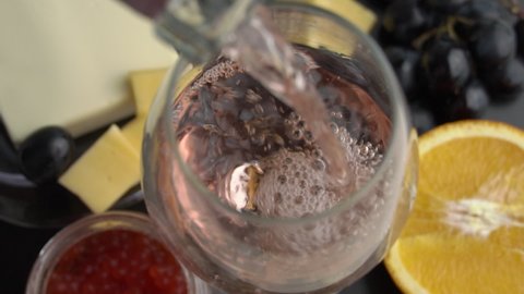 Rose wine is poured into a glass with appetizers in slow mo. Aesthetics of wine with cheese. Alcohol, romance, evening. Cheese, butter, red caviar, grapes, baguette, orange. View from above.