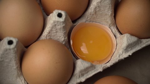 The broken egg lies in the container among the whole eggs. Chicken yolk. Chicken brown fresh raw eggs in a paper box. An egg carton with ten eggs. Lots of fresh raw chicken eggs. Omelette. 4k