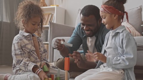 Medium slowmo shot of two cute little girls and their African-American father having fun together while playing dice game sitting on carpet next to couch in cozy living room