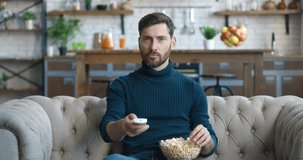 Shocked bearded man watching horror movie on TV and eating popcorn while sitting on the couch at home. Scared millennial male watching terrible news on TV channel while sitting on sofa at living room