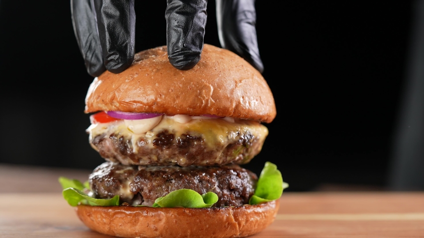 Hand putting top burger bun on a meat double cheeseburger with vegetables with sauce oozing and dripping out on black background. slow motion Royalty-Free Stock Footage #1087069931