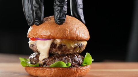 Hand putting top burger bun on a meat double cheeseburger with vegetables with sauce oozing and dripping out on black background. slow motion