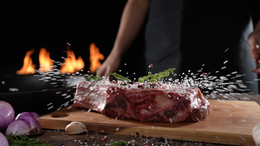 Raw seasoned rib eye steak meat beef dropped on wooden chopping board on a wooden table prepared for cooking with flames in the background. slow motion | Shutterstock HD Video #1087069952