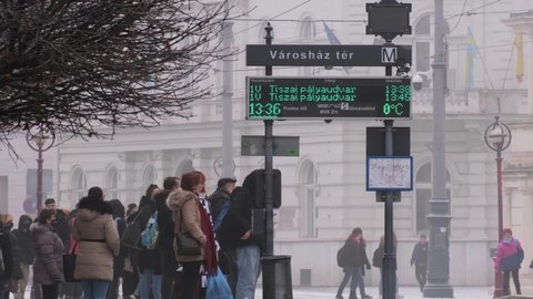 MISKOLC, HUNGARY - JAN 14, 2022: Crowd of people waiting for the tram in a stop. The time is 13:36 and 0°C. Foggy winter afternoon in an east hungarian city. In the foreground pedestrians passing by.