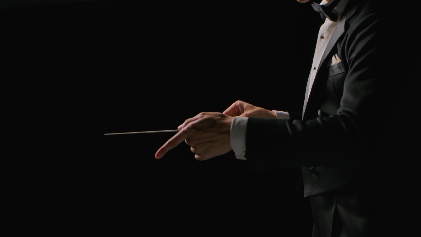 Symphony orchestra conductor wearing suit is directing musicians with movement of baton, isolated on black background. Conducting, directing a musical performance with visible gestures. Close up. Royalty-Free Stock Footage #1087077842