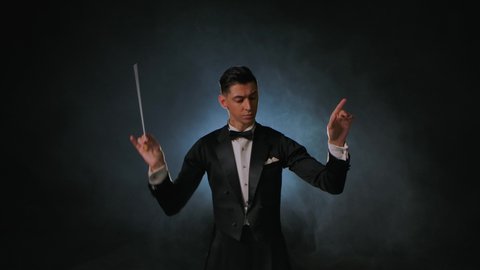An orchestra leader wearing suit using the conducting baton to synchronize the musicians. Maestro is directing musicians with movement of stick on black background with smoke and backlight. Close up.