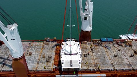 Phuket, Thailand, 22, January, 2022:
A sailing catamaran hanging on slings, lifted by a crane of an international cargo ship, workers install it on props on the deck, view from above