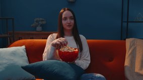 Portrait of attractive woman sitting on sofa alone, eating popcorn and watching interesting tv serial, horror film at home in the living room during evening time. Girl enjoying domestic entertainment