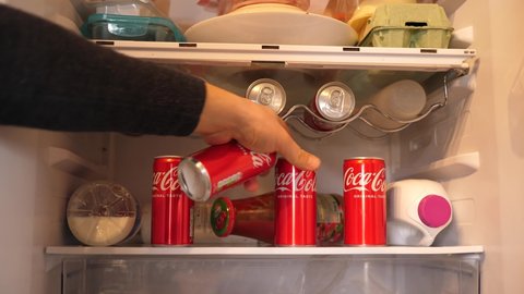 WROCLAW, POLAND - FEB 16, 2022: Person hand takes couple of cans of coca cola carbonated drink from the fridge shelf