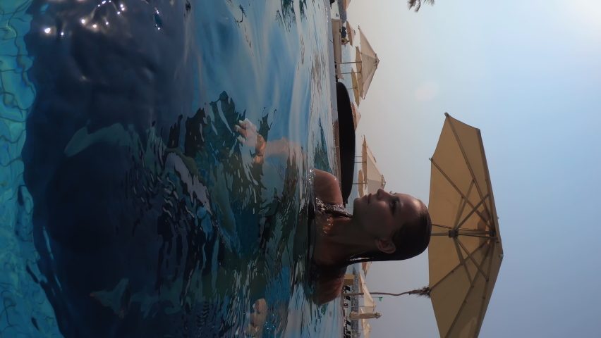 vertical video. Caucasian girl stands in the pool and shows a stop sign with her hands. A European woman in a swimsuit swims. Hot sunny day. Sun umbrella against a clear sky.