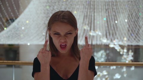 Caucasian girl gestures fuck sign. A beautiful European woman in a black dress on a background of a large crystal chandelier shows her middle finger. Portrait of a cruel lady.