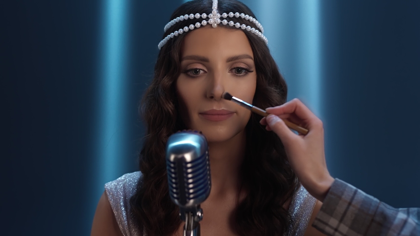 Beautiful Caucasian singer make up on set. artist is being dusted. The brunette stands at the microphone. A blue background with searchlights. The make-up artist's hand corrects the singer's make-up. Royalty-Free Stock Footage #1087086158