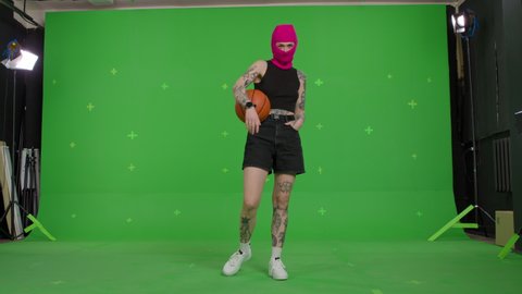 Young woman in pink balaclava with basketball in hands. Hooligan girl in mask looking at camera on green screen background . Chroma key . 4k uhd video