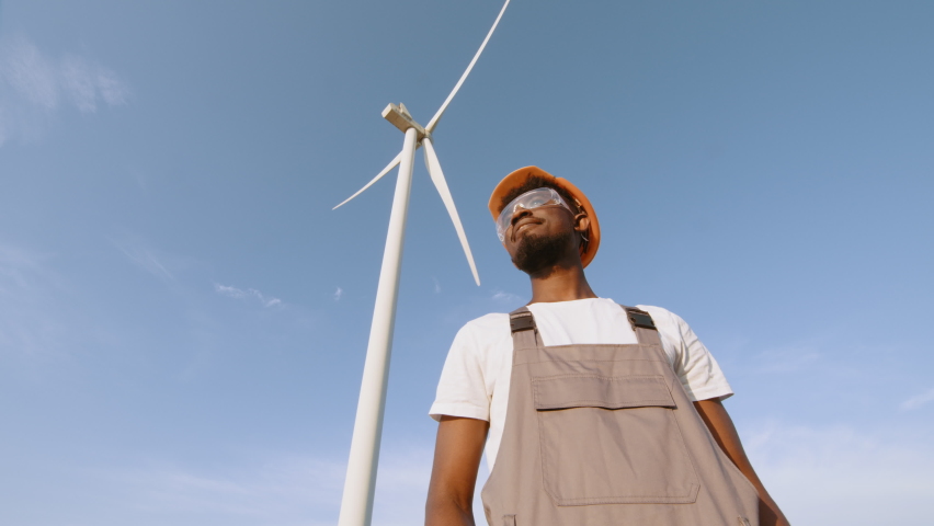 African american technician in helmet, glases and overalls looking at camera while standing on field with wind turbines. Safety electricity and innovation concept. Renewable energy of the future