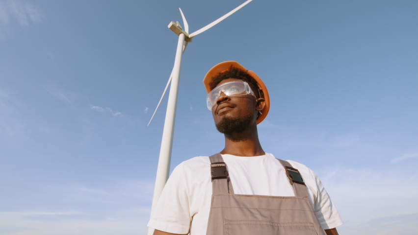 African american technician in helmet, glases and overalls looking at camera while standing on field with wind turbines. Close up man on ecological station. Wind turbines rotating. Renewable energy.
