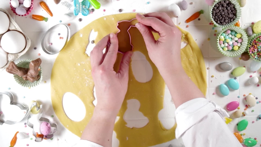 Woman hands kneading dough on table and cooking Easter sugar cookies. Easter baking background with colorful chocolates, candy eggs, baking utencils, ingredients, Top view