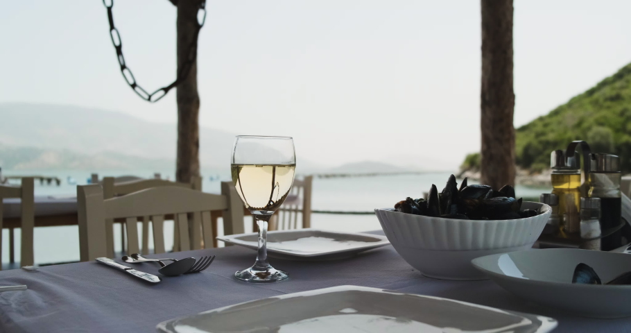 outdoor restaurant, on table filled with a glass of white wine, mussels, table setting. View of the mountains and the sea.