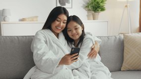 Young carefree asiana mother web surfing online on smartphone, embracing with her little daughter on couch at home, zoom in shot, slow motion