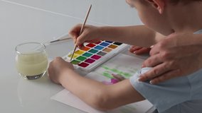 Close-up of a little boy drawing with paints and a brush with his mother at a white table. Boy learns to draw with watercolor paints together with a caring mother. 4k high quality slow motion video