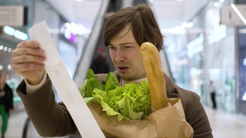 Man gets shocked looking at check after shopping in supermarket. Young consumer holds paper bag with fresh products standing in mall close view
