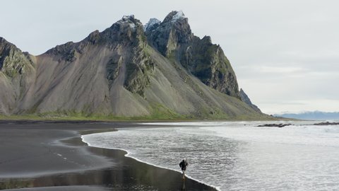 Drone Shot Of Man On Black Sand Beach Walking And Running Along The Tide Coming In With Vestrahorn Mountain In The Background, Iceland