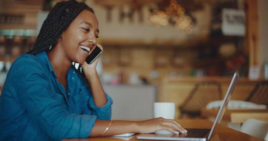 Beautiful successful black woman working remotely in coffee shop on her laptop and mobile phone, sucess and diversity, future of remote work, young successful african american entrepreneur