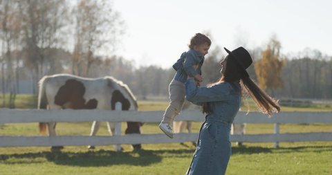 Mother is spinning her little son around on a sunny day with horses at background.  Family photo shoot with with kids. Sunny, bright day. 4k  60p to 23.976 slow motion. medium shoot.