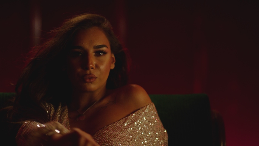 4K Beautiful attractive woman sitting on sofa dancing , flirting , posing and looking into camera with shiny dress . Fashion advertisement concept . Shot on ARRI ALEXA cinema camera in slow motion