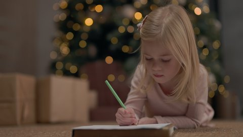 Little Girl Of Four Years With Blond Long Hair. Behind It Christmas Tree Sparkles With Garlands. Girl Draws. She Is On Floor Near Christmas Tree. Near Christmas Tree Gifts.