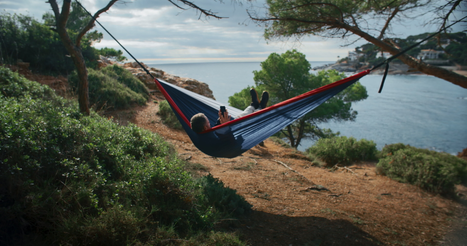 Man chill and relax in camping hammock between two trees in forest on beach shoreline. Relaxing summer or spring vacation. Summertime holiday. scroll through social media on smartphone Royalty-Free Stock Footage #1087118249