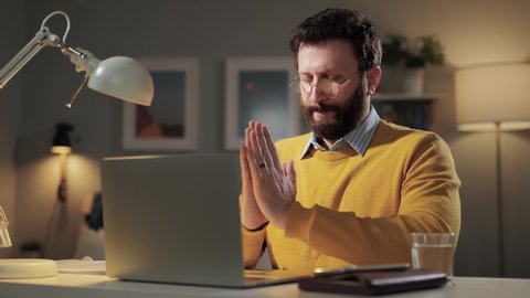 Meditation. Peaceful bearded man sits at desk with his palms joined in front of computer and meditates, breathes deeply and gets to work