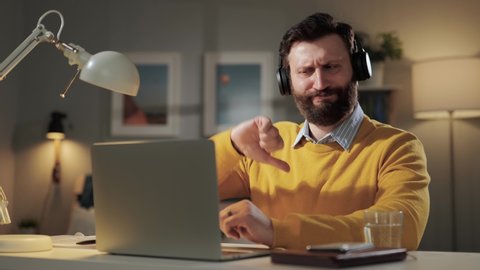 Man thumbs down, disagree emotion. Serious displeased bearded man with headphones in home office looking at computer webcam and shows his hand with thumb down