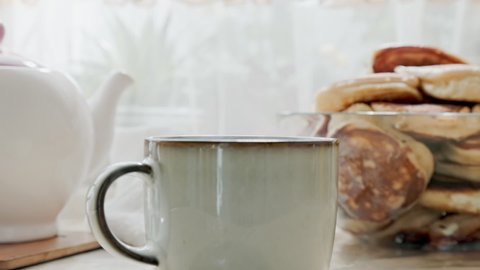 Close up of ceramic mug with hot tea. In the background there is a porcelain white teapot and a bowl of pancakes. Home tea party.