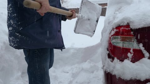 A man in a jacket and jeans is shoveling snow off a red car. The car got snowed in during the blizzard. Snow falls from the sky